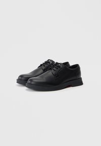 Chaussures en cuir noir avec une surface texturée, bout arrondi, design à lacets et une semelle en caoutchouc avec un discret accent rouge au niveau du talon.