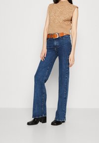 Nainen, jolla on päällään hihaton beige neulepaita, korkeavyötäröiset siniset farkut ruskealla vyöllä ja mustat korkeat nilkkasaappaat, seisoo valkoista seinää vasten.