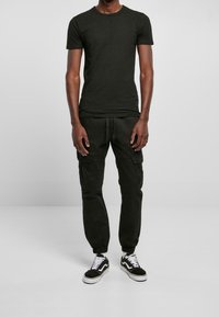 Uomo che indossa una t-shirt nera aderente, pantaloni cargo jogger neri e sneaker bianco-neri, in piedi davanti a uno sfondo semplice.