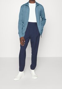 Maglione azzurro chiaro con zip e lavorazione a maglia testurizzata, indossato sopra una t-shirt bianca. Pantaloni blu scuro abbinati a sneaker bianche.