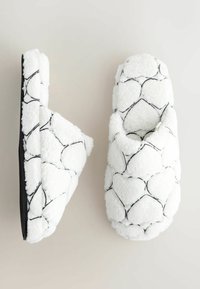 Fuzzy hvide tøfler med et tekstureret mønster af sorte konturer, der ligner hjerter. Blødt materiale og åben bagdesign for nem påtagning.