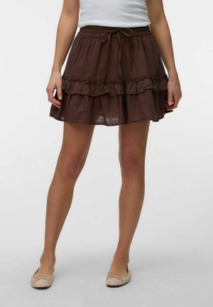 Femme portant une mini-jupe marron à volants superposés avec une taille à cordon, associée à des ballerines beige et un top blanc sans manches.