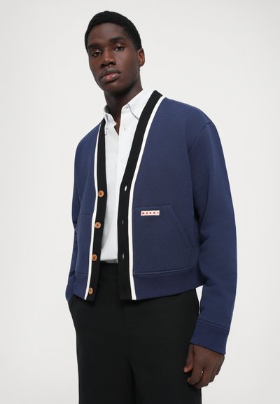 Navy blue cardigan with a black and white trim, wooden buttons, and a front pocket featuring a red label. Soft, textured fabric.