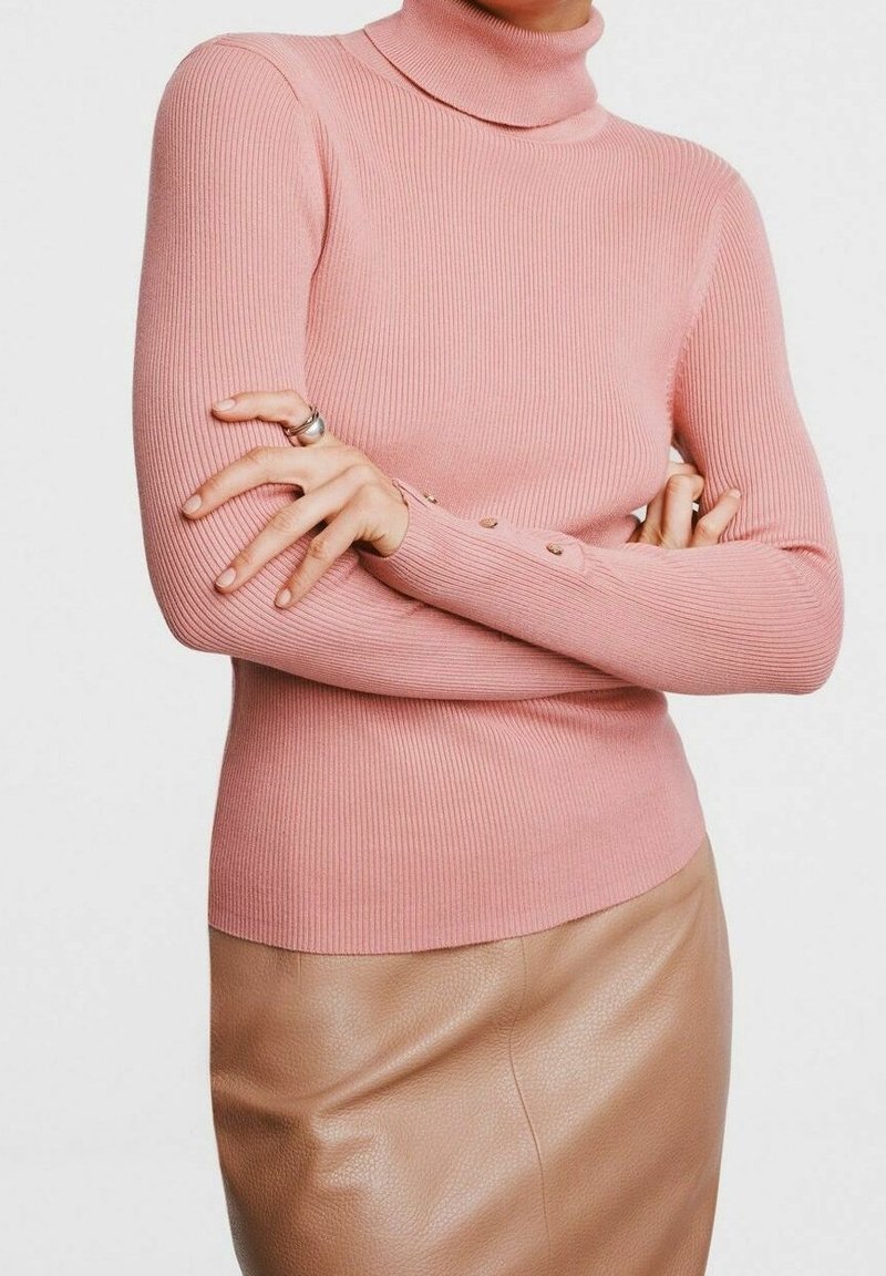 Personne portant un pull à col roulé rose côtelé avec des détails de boutons sur les manches, croisé sur une jupe en cuir beige.