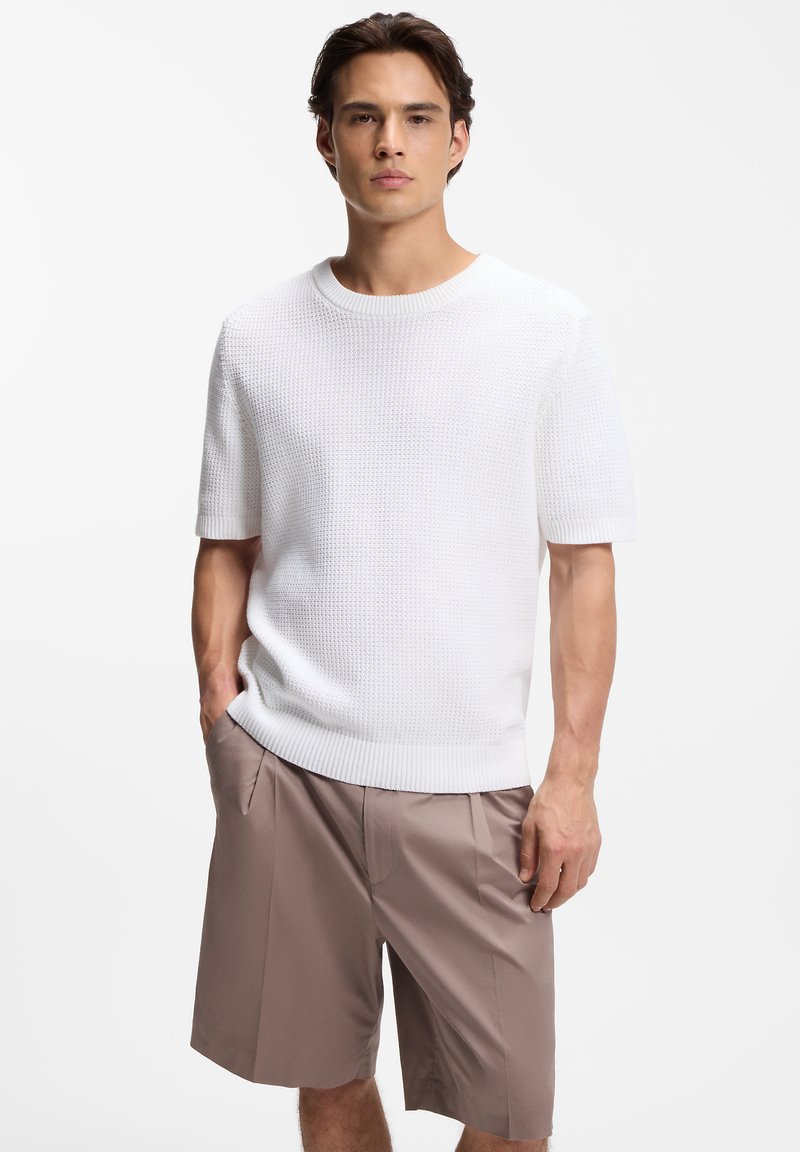 Jeune homme aux cheveux bruns courts portant un pull blanc texturé à manches courtes et un short beige ajusté arrivant au genou.