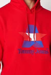 Felpa rossa con cappuccio caratterizzata da una grande grafica a stella in blu e rosso, con linee orizzontali. Logo 'Tommy Jeans' ricamato in blu sotto la grafica.