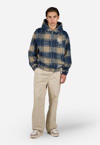 Hettejakke med glidelås i rutete mønster i blått og beige fleece, brukt over beige bukser med lommer og hvite sneakers. Har brodert logo.