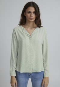 Chemise à manches longues vert menthe avec des pois blancs, en tissu doux et avec un col en V, portée avec un jean bleu.