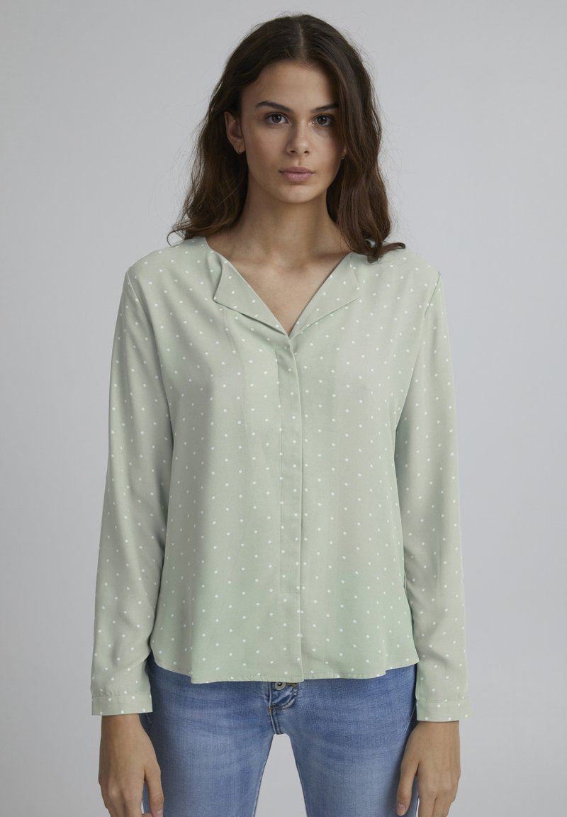 Chemise à manches longues vert menthe avec des pois blancs, en tissu doux et avec un col en V, portée avec un jean bleu.