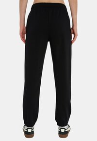 Pantaloni della tuta neri con vita elastica e polsini alle caviglie, realizzati in tessuto morbido, visti da dietro, abbinati a sneakers con dettagli bianchi.