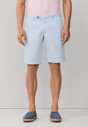 Hombre con pantalones cortos azul claro hasta la rodilla, camisa rosa y zapatos azules sin cordones, de pie contra un fondo sencillo.