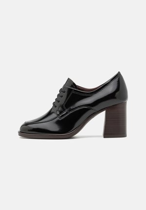 Chaussures à talons en cuir verni noir avec un design à lacets, bout rond et un large talon en bois. Texture lisse et silhouette élégante.