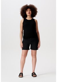 Zwarte tanktop, zwarte denim shorts, platte zwarte sandalen. Model met krullend haar staat rechtop tegen een neutrale achtergrond.