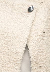 Gebroken-witte, gestructureerde boucle jas met grote glanzende zilveren knoop, gedragen over een geribbeld wit stoffen kledingstuk eronder.