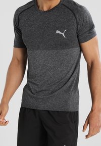 Šedé sportovní tričko s texturovanou vrchní částí, krátkými rukávy a bílým logem Puma na hrudi. Hladká látka, vypasovaný střih.