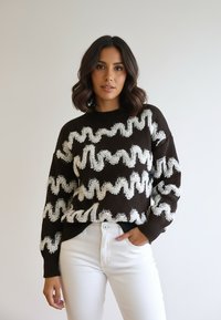 Brązowy sweter z dzianiny z białym falistym wzorem, długimi rękawami i okrągłym dekoltem, w połączeniu z białymi spodniami. Gładka tekstura.