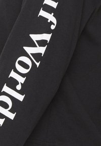 Sort langærmet skjorte med hvid tekst trykt lodret på ærmet. Fremstillet af en bomuldsblanding med en glat tekstur.