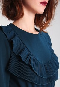 Tealfärgad blus med rund hals och framträdande veckade volanger längs framsidan, tillverkad av mjukt tyg med en lätt glans.