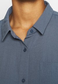 Chemise à manches courtes, boutonnée, en bleu foncé avec une texture tissée. Elle comporte un col, deux boutons près de l'encolure, et une poche poitrine à gauche.