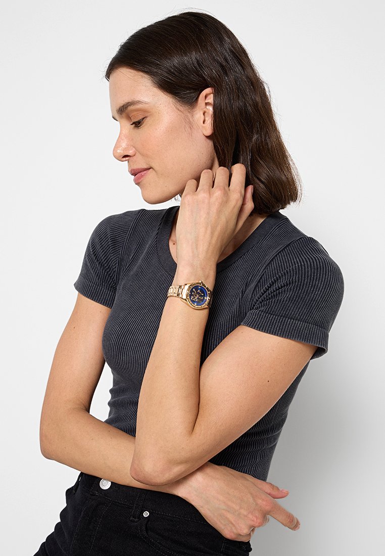 Złoty zegarek na nadgarstek z granatową tarczą, metalową bransoletą i okrągłą kopertą. Noszony na nadgarstku, z teksturowanym czarnym wierzchem.