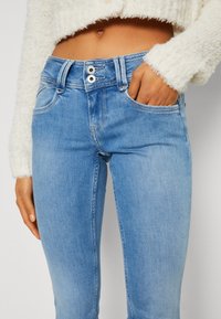 Žena v světlomodrých džínách se dvojitým zapínáním a krémovým chlupatým svetrem, s jednou rukou v kapse, odhalující břicho.