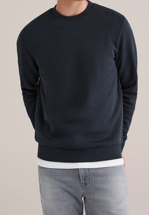 Homme portant un sweat-shirt col rond bleu marine foncé superposé sur une chemise blanche et un jean gris clair, debout devant un fond uni.