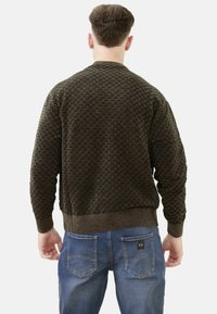 Maglione marrone textured con scollo rotondo e polsini a costine, abbinato a jeans blu, con un logo visibile sulla vita.