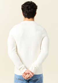 Weißer Strickpullover mit strukturiertem Muster und horizontalen Streifen, Rundhalsausschnitt und gerippten Bündchen, kombiniert mit hellblauen Jeans.