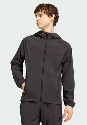 Junger Mann in schwarzer Kapuzenjacke und passender Hose, Hände in den Taschen, schaut nach links vor einem schlichten Hintergrund.