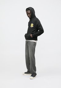 Felpa nera con cappuccio e design grafico giallo, abbinata a jeans grigi a gamba dritta e sneakers nere con suole bianche.