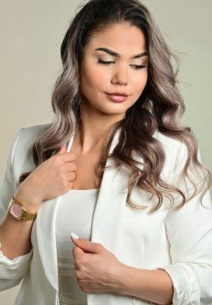 White blazer over a fitted top, featuring long, wavy hair. Gold watch with a pink face and textured band. Light, neutral background.