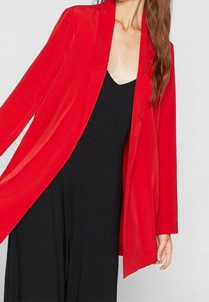 Femme portant un long blazer rouge par-dessus une robe noire sans manches à col en V, avec de longs cheveux bruns tombant sur une épaule.