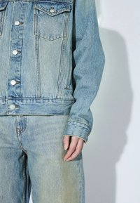 Veste en jean bleu clair avec des boutons argentés assortie à un jean en denim bleu clair assorti, montrant le torse et la main droite d'une personne.