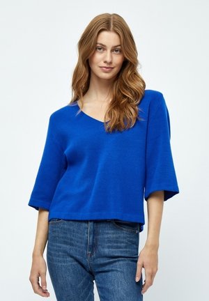 Kombinierter, blauer, gestrickter Pullover mit V-Ausschnitt und Dreiviertelärmeln; getragen mit blauen Jeans, glatte Struktur, keine sichtbaren Muster oder Akzente.