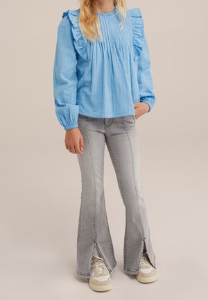 Dziecko ubrane w jasnoniebieską bluzkę z długim rękawem i falbankami oraz szare dzwony z przednimi rozcięciami, do tego białe trampki.