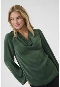 Blouse en satin vert avec un décolleté drapé et des manches longues, présentant une texture lisse et un doux éclat. Des pantalons noirs complètent la tenue.