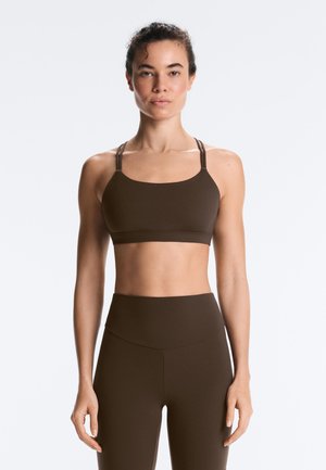 Kobieta w brązowym sportowym staniku i legginsach z wysokim stanem, stoi prosto na tle jednolitego białego tła.