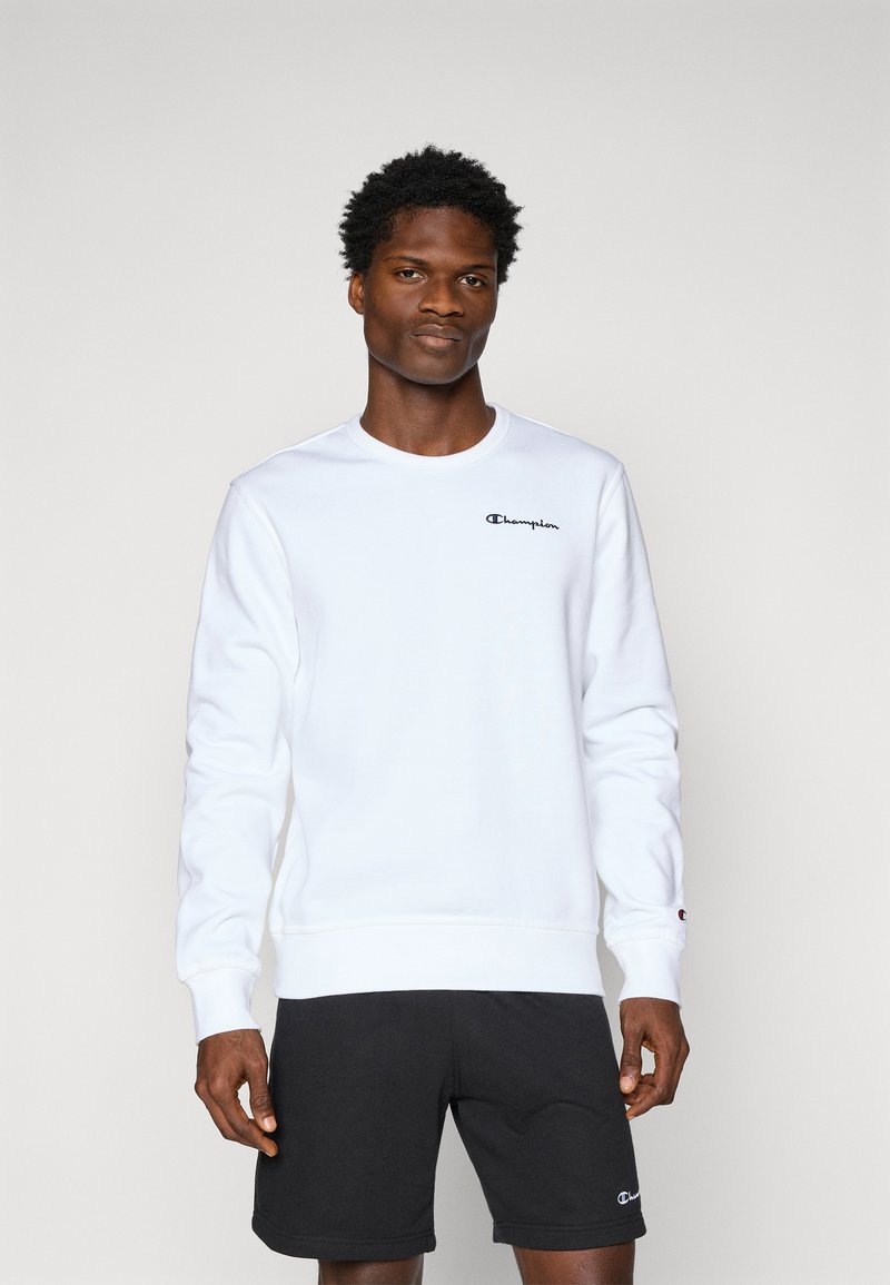 Sweatshirt blanc avec des poignets et un ourlet côtelés, arborant un petit logo Champion noir sur la poitrine. Porté avec un short de sport noir.