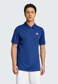 Blå pikétrøye laget av pustende stoff, med en tre-knapps knappestolpe og en hvit logo på venstre bryst. Glatt tekstur, kortermet.