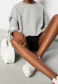 Sudadera gris con un logo dorado de Nike, combinada con pantalones cortos negros. Bolso blanco y zapatillas de color blanco sucio. Textura de piel suave y una pose casual.