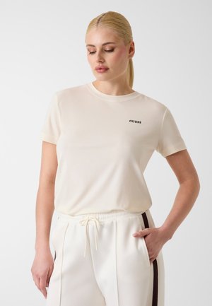 Femme blonde portant un t-shirt Guess crème et un pantalon blanc à cordon avec des bandes noires sur les côtés, posant avec une main dans la poche.