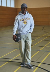 Svetlo modra kapuca s logotipom "Los Angeles Lakers", udobne svetlo zeleni hlače, črna košarka v roki in črne športne superge.