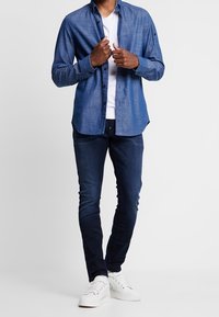 Hombre vestido con una camisa de botones azul sobre una camiseta blanca, jeans ajustados de mezclilla oscura y zapatillas blancas, ajustándose el cuello de la camisa.