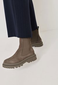 Bruna mocka ankboots med elastiska sidopaneler, kraftiga gummisulor och texturerad detaljering. Bärs med marinblå randiga byxor.