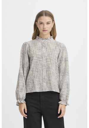 Giovane donna che indossa una camicetta a maniche lunghe, con bottoni, a quadri, con colletto e polsini arricciati, abbinata a pantaloni neri, in piedi, eretta.