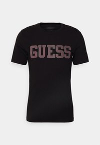 Černé bavlněné tričko s krátk�ým rukávem, na kterém je přes hrudník umístěn nápis "GUESS" ve svislých pruhovaných červených a bílých písmenech.