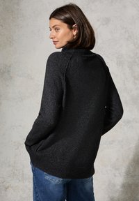 Maglione nero lavorato a maglia con colletto alto, finitura testurizzata e orlo a coste. Indossato sopra dei jeans blu, mostra una vestibilità rilassata e maniche lunghe.