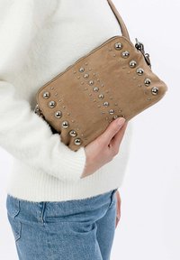 Tasche aus beigem Stoff mit silbernen Nieten in gepunktetem Muster, mit Reißverschluss und einem kurzen Tragegurt, gehalten gegen einen weißen Pullover.