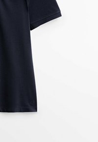 Tissu de polo bleu marine foncé à manches courtes avec détail de couture sur fond blanc, montrant l'avant-bras et le côté.