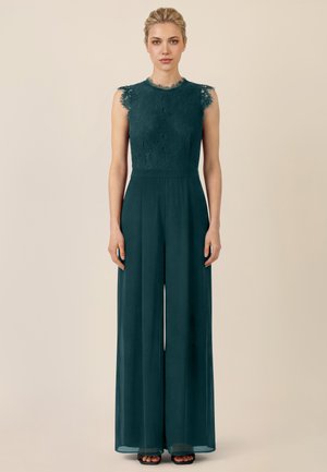 Woman standing facing forward wearing sleeveless dark green lace top jumpsuit with wide-leg pants and black open-toe heels on beige background.